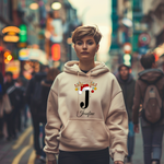 Auf dem Bild ist eine junge Frau zu sehen, die einen weißen Hoodie trägt. Auf dem Hoodie ist ein festliches Design mit einem großen schwarzen Buchstaben sowie der Namen „Josefine“ passend zum Buchstaben, in eleganter Schrift gedruckt. Über dem Buchstaben sind ein Rentiergeweih, ein Weihnachtsmann-Hut und bunte Kugeln als weihnachtliche Dekoration angebracht. Die Frau steht in einer belebten, unscharf abgebildeten Stadtstraße mit Lichtern und Menschen im Hintergrund, mit einer winterlichen, Atmosphäre. 