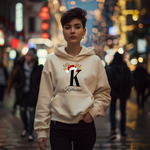 Auf dem Bild ist eine junge Frau zu sehen, die einen weißen Hoodie trägt. Auf dem Hoodie ist ein festliches Design mit einem großen schwarzen Buchstaben sowie der Namen „Katharina“ passend zum Buchstaben, in eleganter Schrift gedruckt. Über dem Buchstaben sind ein Rentiergeweih, ein Weihnachtsmann-Hut und bunte Kugeln als weihnachtliche Dekoration angebracht. Die Frau steht in einer belebten, unscharf abgebildeten Stadtstraße mit Lichtern und Menschen im Hintergrund, mit einer winterlichen, Atmosphäre. 