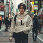 Auf dem Bild ist eine junge Frau zu sehen, die einen weißen Hoodie trägt. Auf dem Hoodie ist ein festliches Design mit einem großen schwarzen Buchstaben sowie der Namen „Leandra“ passend zum Buchstaben, in eleganter Schrift gedruckt. Über dem Buchstaben sind ein Rentiergeweih, ein Weihnachtsmann-Hut und bunte Kugeln als weihnachtliche Dekoration angebracht. Die Frau steht in einer belebten, unscharf abgebildeten Stadtstraße mit Lichtern und Menschen im Hintergrund, mit einer winterlichen, Atmosphäre. 