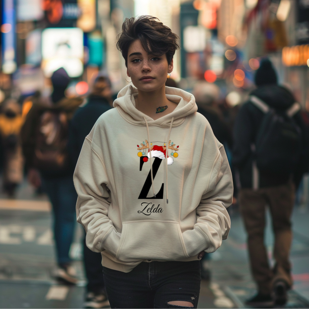 Auf dem Bild ist eine junge Frau zu sehen, die einen weißen Hoodie trägt. Auf dem Hoodie ist ein festliches Design mit einem großen schwarzen Buchstaben sowie der Namen „Zelda“ passend zum Buchstaben, in eleganter Schrift gedruckt. Über dem Buchstaben sind ein Rentiergeweih, ein Weihnachtsmann-Hut und bunte Kugeln als weihnachtliche Dekoration angebracht. Die Frau steht in einer belebten, unscharf abgebildeten Stadtstraße mit Lichtern und Menschen im Hintergrund, mit einer winterlichen, Atmosphäre. 