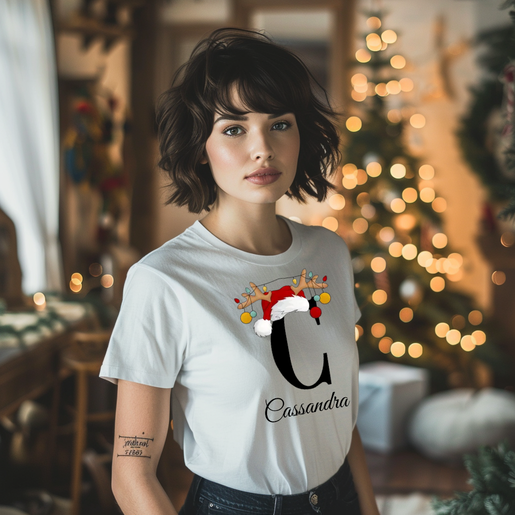 Ein weißes T-Shirt mit einem weihnachtlichen Design, das den Buchstaben 'C' zeigt. Der Buchstabe ist in schwarz gehalten und mit einem Weihnachtsmann-Hut sowie Rentiergeweihen verziert, an denen bunte Kugeln hängen. Unter dem Buchstaben ist der personalisierte Name 'Cassandra' in eleganter, geschwungener Schrift zu sehen. Das T-Shirt strahlt eine festliche und personalisierte Stimmung aus und eignet sich perfekt für die Weihnachtszeit.