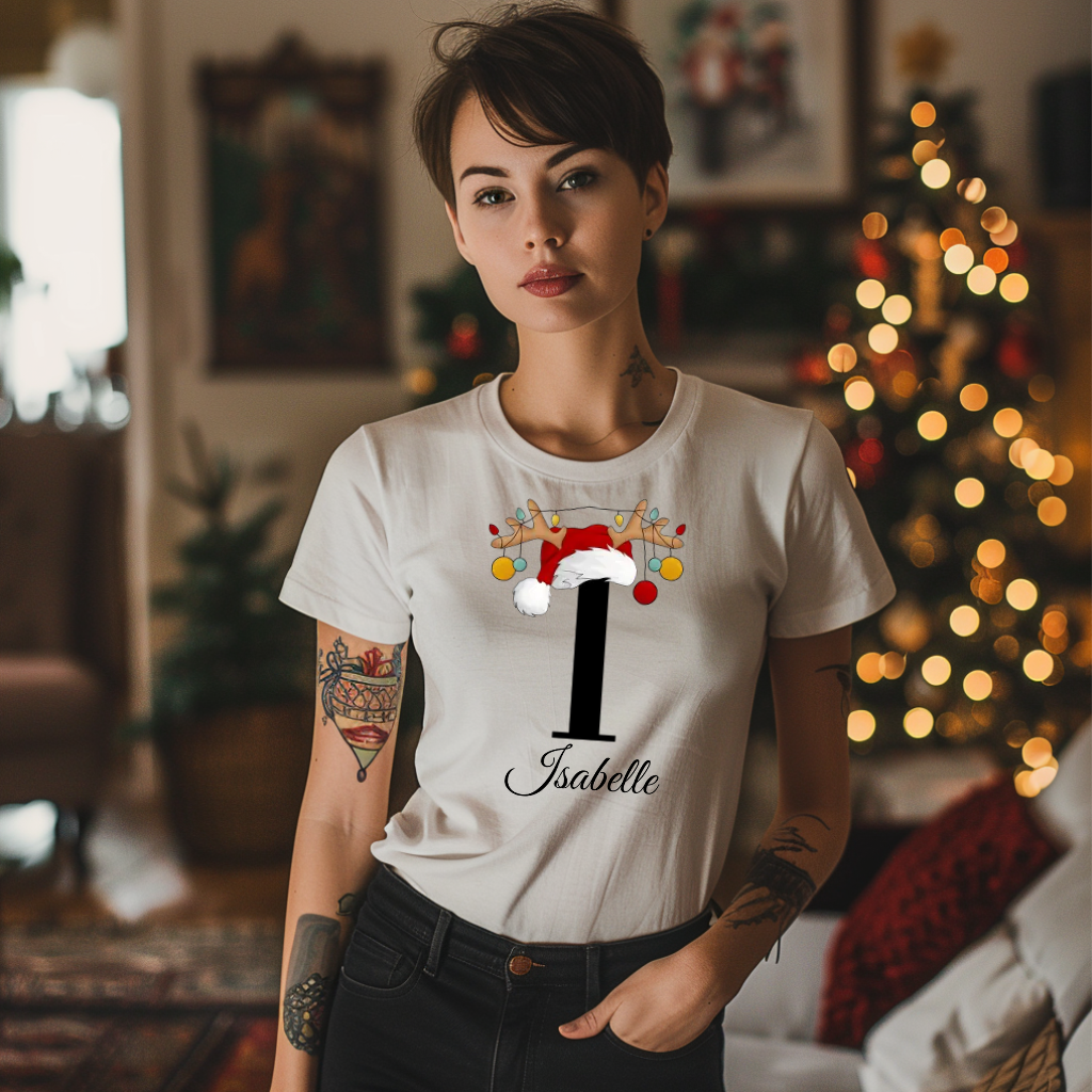 Ein weißes T-Shirt mit einem festlichen Design. Der Buchstabe 'I' ist in Schwarz dargestellt und wird von einem Weihnachtsmann-Hut und Rentiergeweihen geschmückt, an denen bunte Kugeln hängen. Darunter ist der Name 'Isabelle' in eleganter, geschwungener Schrift platziert. Das T-Shirt verbreitet eine personalisierte, weihnachtliche Atmosphäre und ist perfekt für die festliche Jahreszeit geeignet.