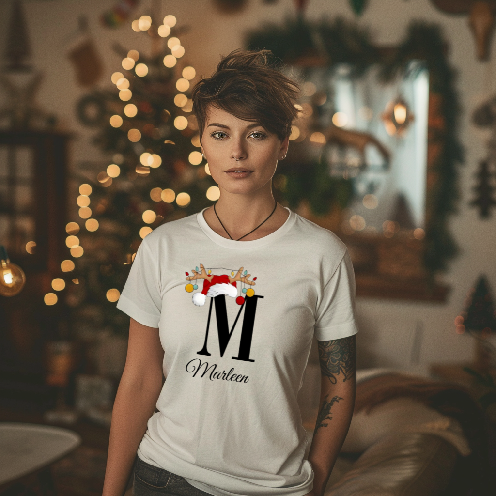 Ein weißes T-Shirt mit einem weihnachtlichen Design, das den Buchstaben 'M' zeigt, verziert mit einem Weihnachtsmann-Hut und Rentiergeweihen, an denen bunte Kugeln hängen. Unter dem Buchstaben steht der Name 'Marleen' in eleganter, geschwungener Schrift.