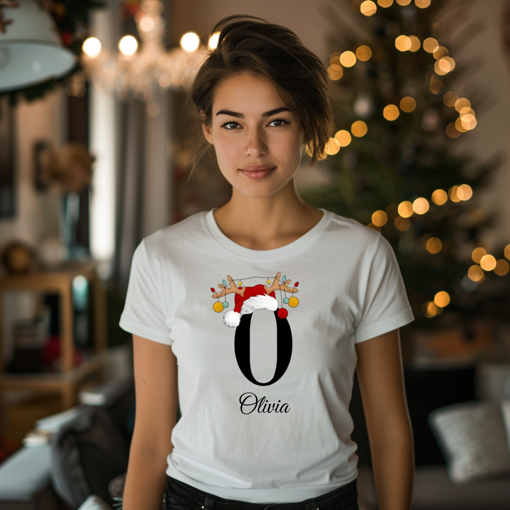 Ein weißes T-Shirt, das den Buchstaben 'O' in Schwarz zeigt. Der Buchstabe ist mit einem Weihnachtsmann-Hut und Rentiergeweihen geschmückt, an denen bunte Kugeln hängen. Darunter ist der Name 'Olivia' in einer eleganten, geschwungenen Schrift abgebildet.