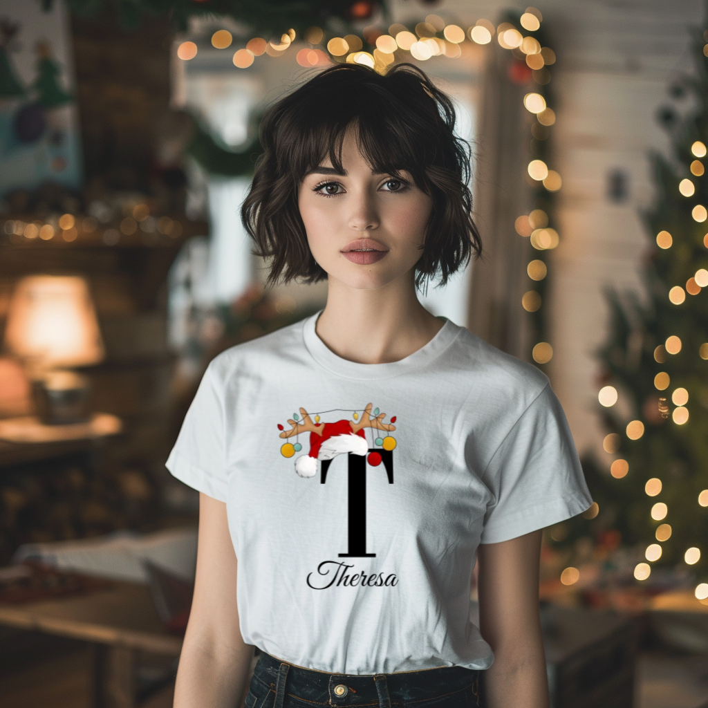 Ein schlichtes weißes T-Shirt, in der Mitte verziert mit einem großen schwarzen Buchstaben 'T', geschmückt mit einem Weihnachtsmann-Hut und Rentiergeweihen, an denen bunte Kugeln hängen. Darunter steht in eleganter Schrift der Name 'Theresa'. Perfekt für die festliche Weihnachtszeit.