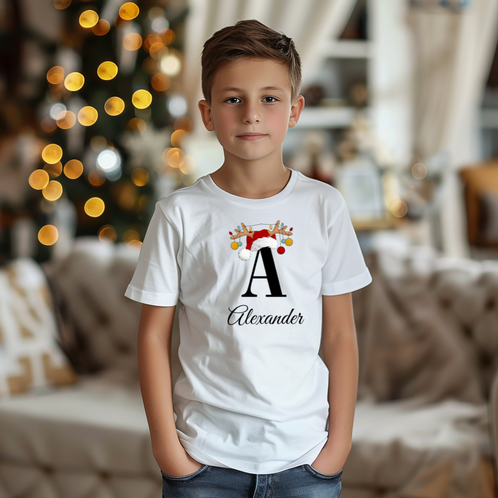 Ein Junge trägt ein weißes T-Shirt mit einem personalisierten Weihnachtsdesign. Der Buchstabe ‚A‘ ist groß auf der Brust zu sehen, verziert mit einem Weihnachtsmann-Hut und Rentiergeweihen mit bunten Kugeln. Darunter steht der Name ‚Alexander‘ in eleganter Schrift. Im Hintergrund strahlt eine gemütliche Weihnachtsatmosphäre mit einem unscharfen Weihnachtsbaum.