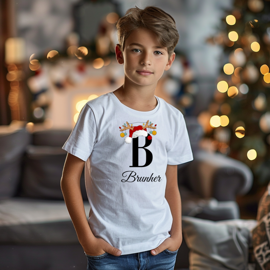 Ein Junge trägt ein weißes T-Shirt mit einem personalisierten Weihnachtsdesign. Der Buchstabe ‚B‘ ist groß auf der Brust zu sehen, verziert mit einem Weihnachtsmann-Hut und Rentiergeweihen, an denen bunte Kugeln hängen. Darunter steht der Name ‚Brunher‘ in eleganter, geschwungener Schrift. Im Hintergrund schafft eine gemütliche Weihnachtsdekoration mit Lichtern eine festliche Atmosphäre.