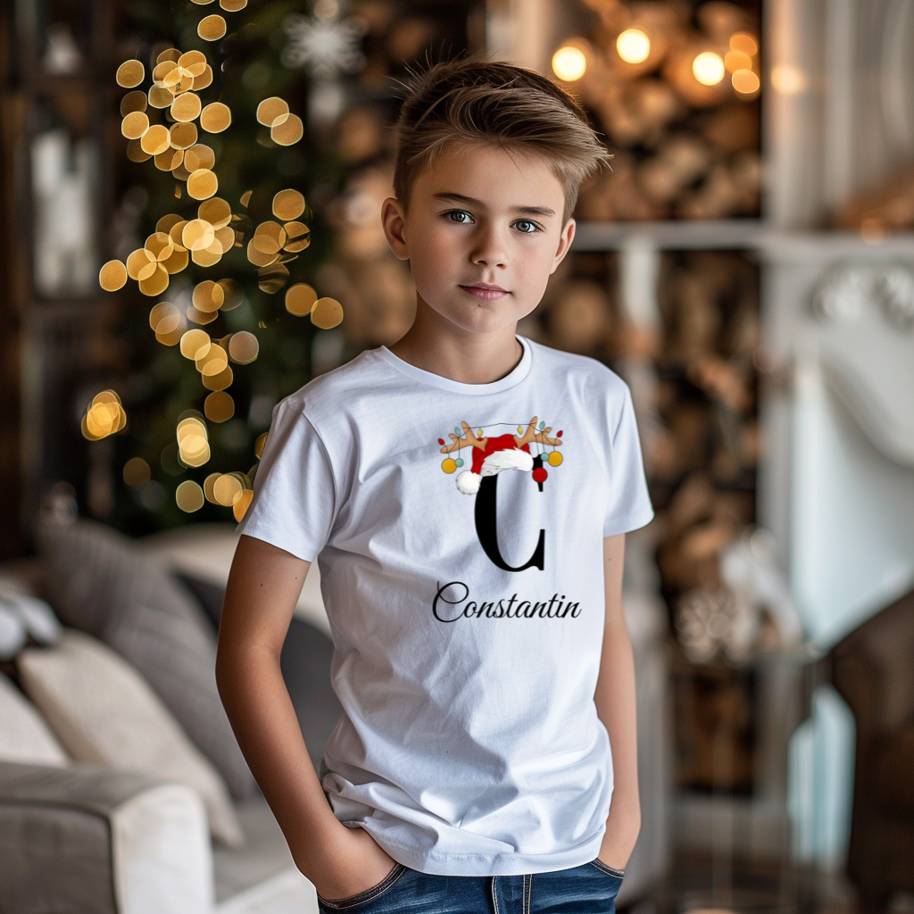 Ein Junge trägt ein weißes T-Shirt mit einem personalisierten Weihnachtsdesign. Der Buchstabe ‚C‘ ist groß auf der Brust zu sehen, verziert mit einem Weihnachtsmann-Hut und Rentiergeweihen, an denen bunte Kugeln hängen. Unter dem Buchstaben steht der Name ‚Constantin‘ in eleganter, geschwungener Schrift. Im Hintergrund sorgt eine weihnachtliche Dekoration mit Lichtern für eine festliche Atmosphäre.