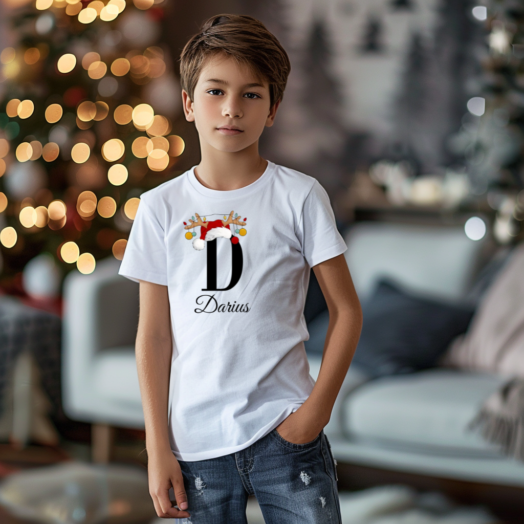 Ein Junge trägt ein weißes T-Shirt mit einem personalisierten Weihnachtsdesign. Der Buchstabe ‚D‘ ist groß auf der Brust zu sehen, verziert mit einem Weihnachtsmann-Hut und Rentiergeweihen, an denen bunte Kugeln hängen. Darunter steht der Name ‚Darius‘ in eleganter, geschwungener Schrift. Im Hintergrund sorgt eine weihnachtliche Dekoration mit Lichtern für eine festliche Atmosphäre.