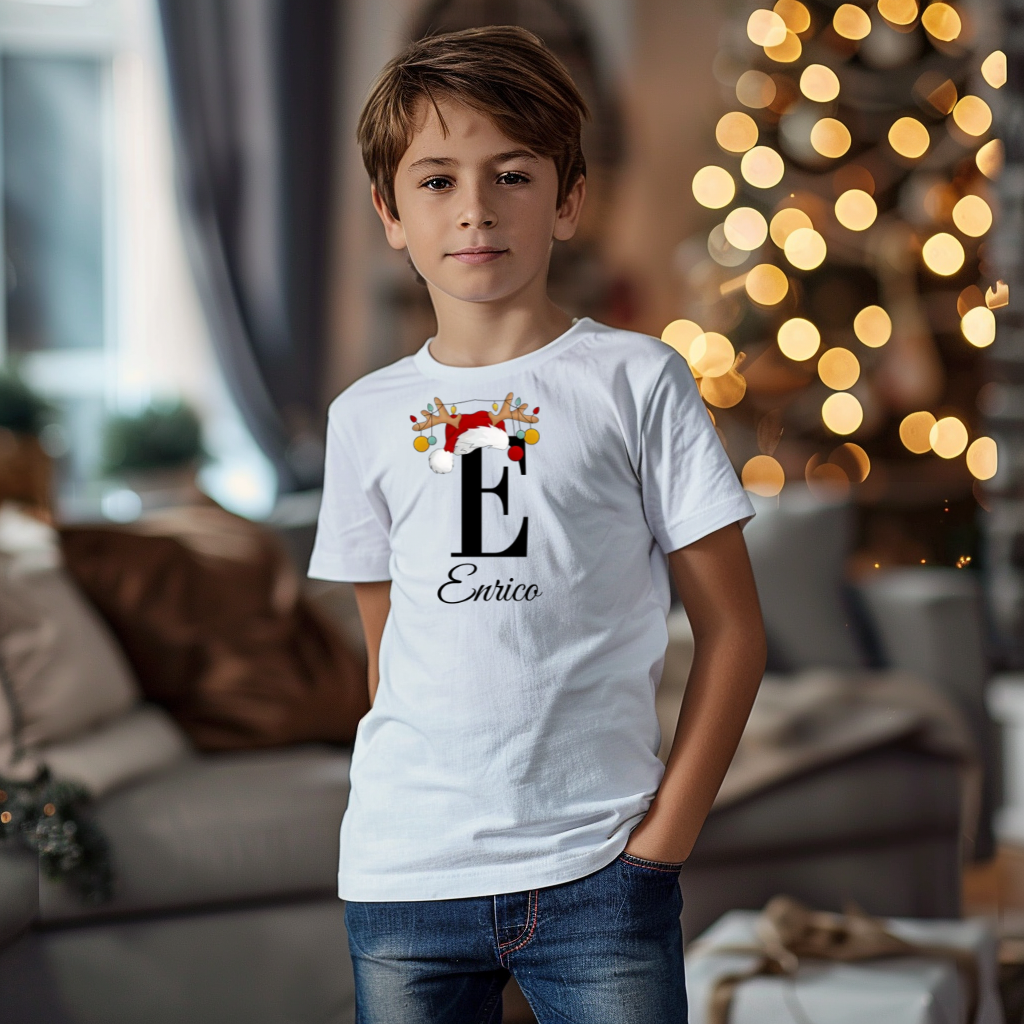 Ein Junge trägt ein weißes T-Shirt mit einem personalisierten Weihnachtsdesign. Der Buchstabe ‚E‘ ist groß auf der Brust zu sehen, verziert mit einem Weihnachtsmann-Hut und Rentiergeweihen, an denen bunte Kugeln hängen. Darunter steht der Name ‚Enrico‘ in eleganter, geschwungener Schrift. Im Hintergrund sorgt eine weihnachtliche Dekoration mit Lichtern für eine festliche Atmosphäre.