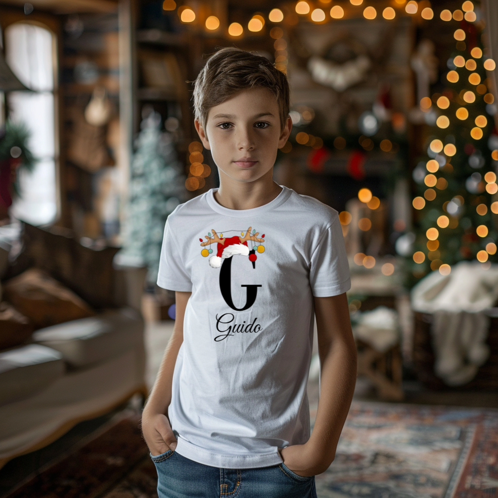 Ein Junge trägt ein weißes T-Shirt mit einem personalisierten Weihnachtsdesign. Der Buchstabe ‚G‘ ist groß auf der Brust zu sehen, verziert mit einem Weihnachtsmann-Hut und Rentiergeweihen, an denen bunte Kugeln hängen. Darunter steht der Name ‚Guido‘ in eleganter, geschwungener Schrift. Im Hintergrund schafft eine weihnachtliche Dekoration mit Lichtern eine festliche Atmosphäre.