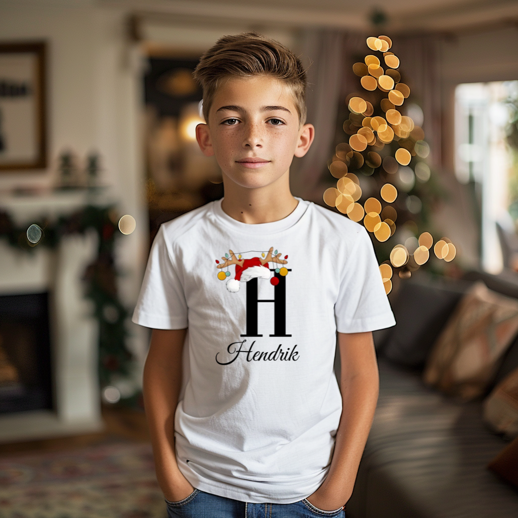 Ein Junge trägt ein weißes T-Shirt mit einem personalisierten Weihnachtsdesign. Der Buchstabe ‚H‘ ist groß auf der Brust zu sehen, verziert mit einem Weihnachtsmann-Hut und Rentiergeweihen, an denen bunte Kugeln hängen. Darunter steht der Name ‚Hendrik‘ in eleganter, geschwungener Schrift. Im Hintergrund sorgt eine weihnachtliche Dekoration mit Lichtern für eine festliche Atmosphäre.