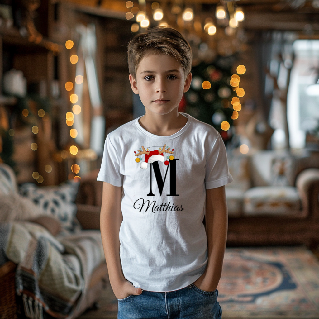 Ein Junge trägt ein weißes T-Shirt mit einem personalisierten Weihnachtsdesign. Der Buchstabe ‚M‘ ist groß auf der Brust zu sehen, verziert mit einem Weihnachtsmann-Hut und Rentiergeweihen, an denen bunte Kugeln hängen. Darunter steht der Name ‚Matthias‘ in eleganter, geschwungener Schrift. Im Hintergrund sorgt eine weihnachtliche Dekoration mit Lichtern für eine festliche Atmosphäre.