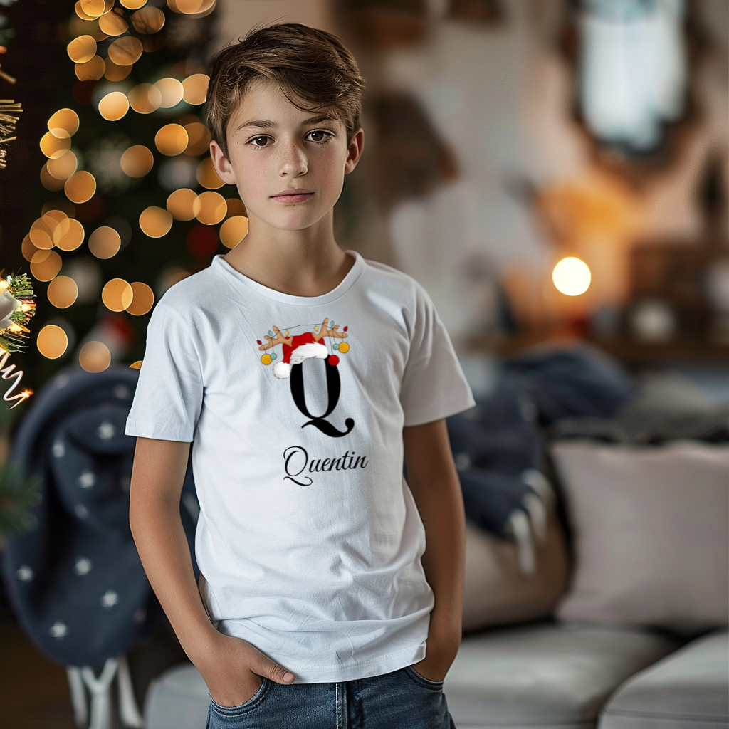 Ein Junge trägt ein weißes T-Shirt mit einem personalisierten Weihnachtsdesign. Der Buchstabe ‚Q‘ ist groß auf der Brust zu sehen, verziert mit einem Weihnachtsmann-Hut und Rentiergeweihen, an denen bunte Kugeln hängen. Darunter steht der Name ‚Quentin‘ in eleganter, geschwungener Schrift. Im Hintergrund sorgt eine weihnachtliche Dekoration mit Lichtern für eine festliche Atmosphäre.