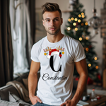 Ein weißes T-Shirt mit einem weihnachtlichen „C“-Design in Schwarz, verziert mit einem Weihnachtsmann-Hut und Rentiergeweihen, an denen bunte Kugeln hängen. Unter dem Buchstaben steht der personalisierte Name „Constantin“ in eleganter, geschwungener Schrift. Der Träger steht in einem weihnachtlich geschmückten Raum mit einem festlichen Weihnachtsbaum im Hintergrund, was eine gemütliche und festliche Atmosphäre schafft. Perfekt für die Weihnachtszeit und individuelle Namensgestaltung.