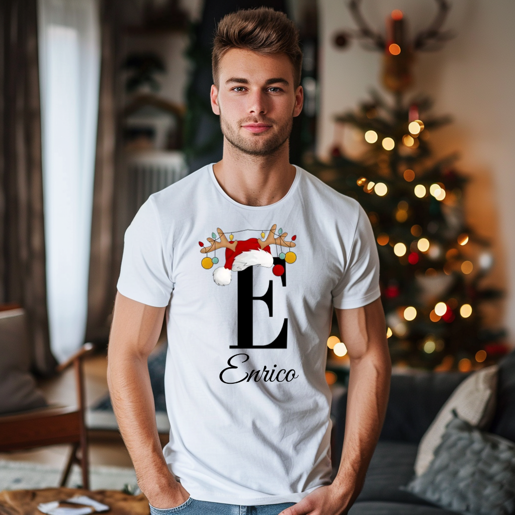 Ein weißes T-Shirt mit einem weihnachtlichen „E“-Design in Schwarz, verziert mit einem Weihnachtsmann-Hut und Rentiergeweihen, an denen bunte Kugeln hängen. Unter dem Buchstaben steht der personalisierte Name „Enrico“ in eleganter, geschwungener Schrift. Der Name kann individuell angepasst werden, während die Schrift unverändert bleibt. Der Träger steht in einem festlich dekorierten Raum mit einem leuchtenden Weihnachtsbaum im Hintergrund, was eine gemütliche und festliche Atmosphäre schafft. Ideal für die 