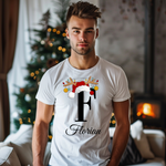 Ein junger Mann steht in einem weihnachtlich dekorierten Raum, der im Hintergrund einen geschmückten Weihnachtsbaum und einen Kamin zeigt. Er trägt ein weißes T-Shirt mit einem festlichen Design. Auf dem T-Shirt ist ein großer Buchstabe "F" abgebildet, der mit einem Weihnachtsmann-Hut und Rentiergeweihen dekoriert ist. An den Geweihen hängen bunte Kugeln, und unter dem Buchstaben steht der Name "Florian" in eleganter, geschwungener Schrift. Das Design vermittelt eine warme und festliche Stimmung, passend zu