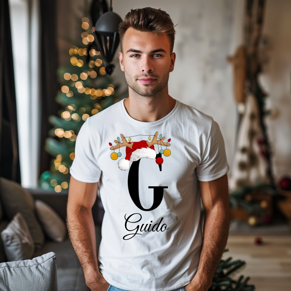 Ein junger Mann steht in einem gemütlich dekorierten Raum, im Hintergrund ist ein geschmückter Weihnachtsbaum und sanfte Beleuchtung zu sehen. Er trägt ein weißes T-Shirt mit einem festlichen Design. Auf dem T-Shirt ist ein großer Buchstabe "G" abgebildet, der mit einem Weihnachtsmann-Hut und Rentiergeweihen dekoriert ist, an denen bunte Weihnachtskugeln hängen. Unter dem Buchstaben steht der Name "Guido" in eleganter, geschwungener Schrift. Das Design vermittelt eine weihnachtliche Stimmung.