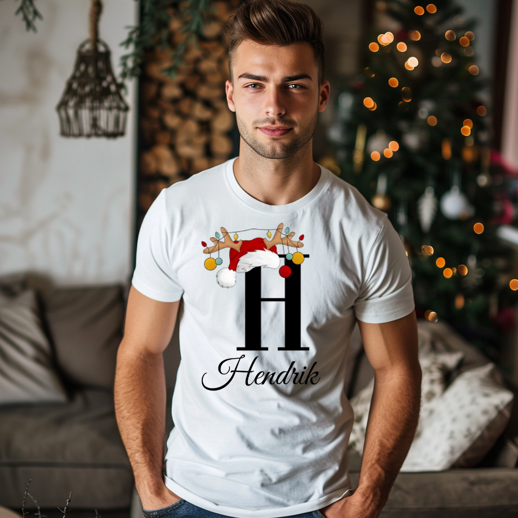 Ein junger Mann steht in einem gemütlich dekorierten Raum, im Hintergrund ist ein Weihnachtsbaum mit Lichtern zu sehen. Er trägt ein weißes T-Shirt mit einem festlichen Design. Auf dem T-Shirt ist ein großer Buchstabe "H" abgebildet, der mit einem Weihnachtsmann-Hut und Rentiergeweihen dekoriert ist, an denen bunte Kugeln hängen. Unter dem Buchstaben steht der Name "Hendrik" in eleganter, geschwungener Schrift. Das Design vermittelt eine weihnachtliche und personalisierte Stimmung.