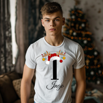 Ein junger Mann steht in einem festlich dekorierten Raum, im Hintergrund ist ein geschmückter Weihnachtsbaum mit Lichtern zu sehen. Er trägt ein weißes T-Shirt mit einem weihnachtlichen Design. Auf dem T-Shirt ist ein großer Buchstabe "I" abgebildet, der mit einem Weihnachtsmann-Hut und Rentiergeweihen dekoriert ist, an denen bunte Kugeln hängen. Unter dem Buchstaben steht der Name "Ingo" in eleganter, geschwungener Schrift. Das Design strahlt eine festliche und personalisierte Stimmung aus.