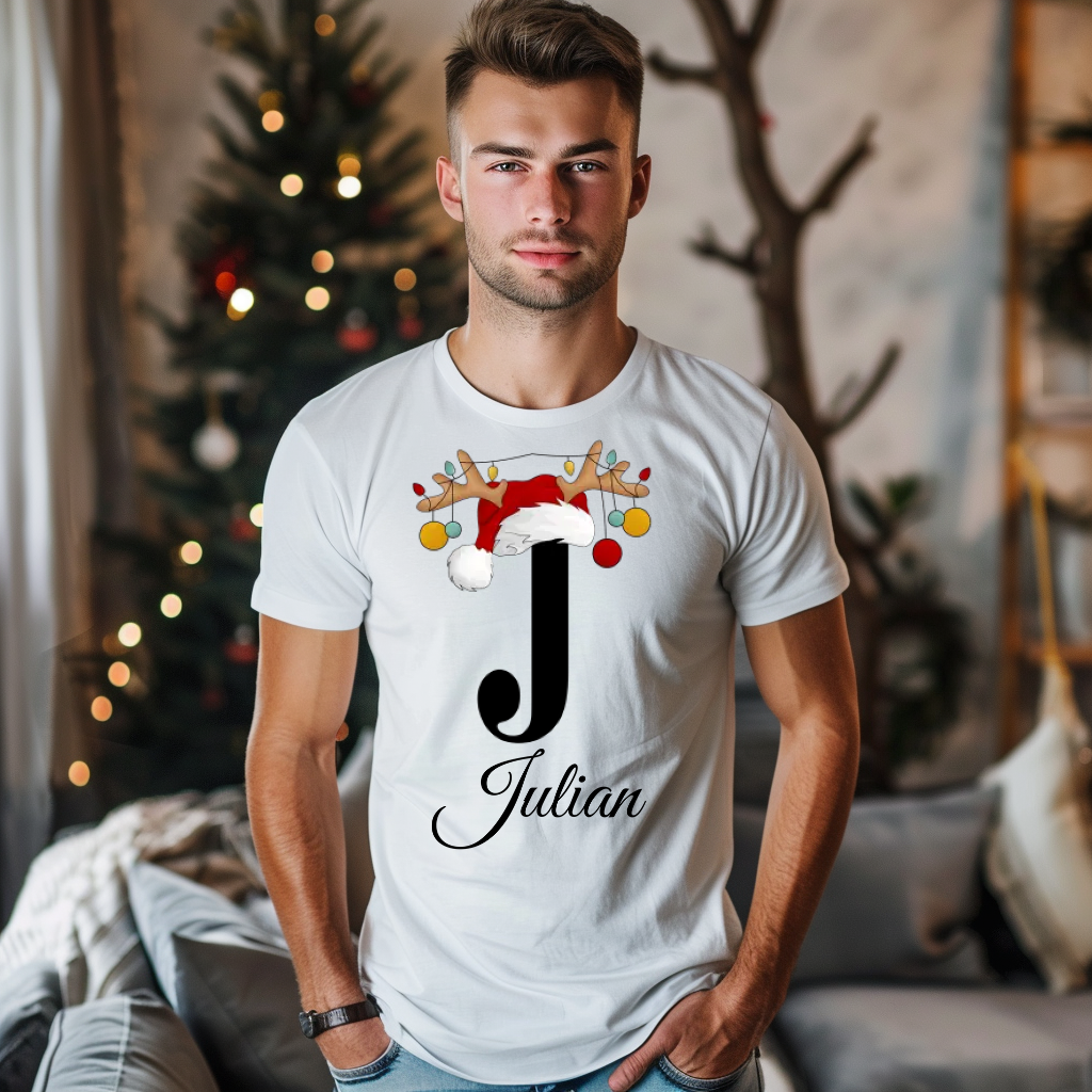 Ein junger Mann steht in einem gemütlichen, weihnachtlich dekorierten Raum, im Hintergrund ist ein geschmückter Weihnachtsbaum zu sehen. Er trägt ein weißes T-Shirt mit einem festlichen Design. Auf dem T-Shirt ist ein großer Buchstabe "J" abgebildet, der mit einem Weihnachtsmann-Hut und Rentiergeweihen dekoriert ist, an denen bunte Kugeln hängen. Unter dem Buchstaben steht der Name "Julian" in eleganter, geschwungener Schrift. Das Design vermittelt eine personalisierte und weihnachtliche Stimmung.