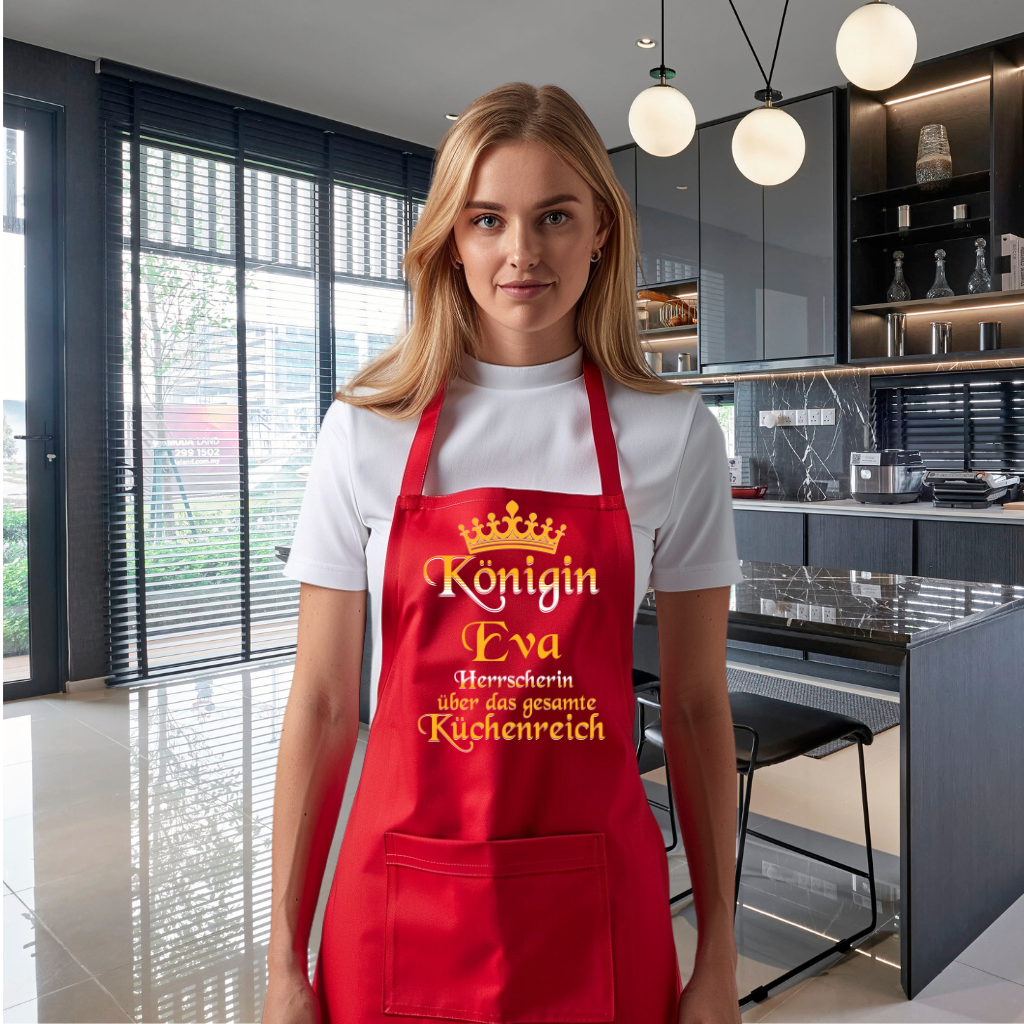 Eine Frau trägt eine rote Grillschürze mit einer goldenen Krone und dem personalisierten Schriftzug 'Königin [Name], Herrscherin über das gesamte Küchenreich' in eleganter Schrift. Die Schürze hat eine praktische Tasche auf der Vorderseite. Im Hintergrund ist eine moderne Küche mit viel natürlichem Licht und eleganten schwarzen Schränken zu sehen.